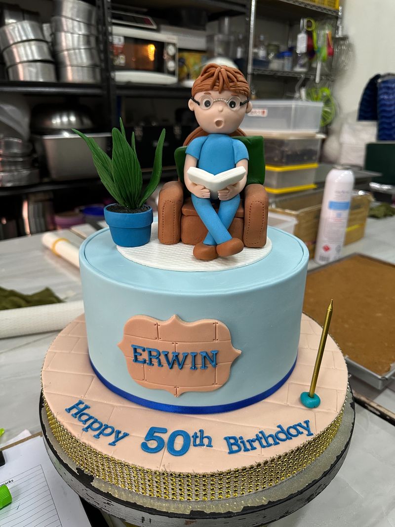 Light-blue 50th birthday cake topped with a figurine reading in an armchair beside a potted plant