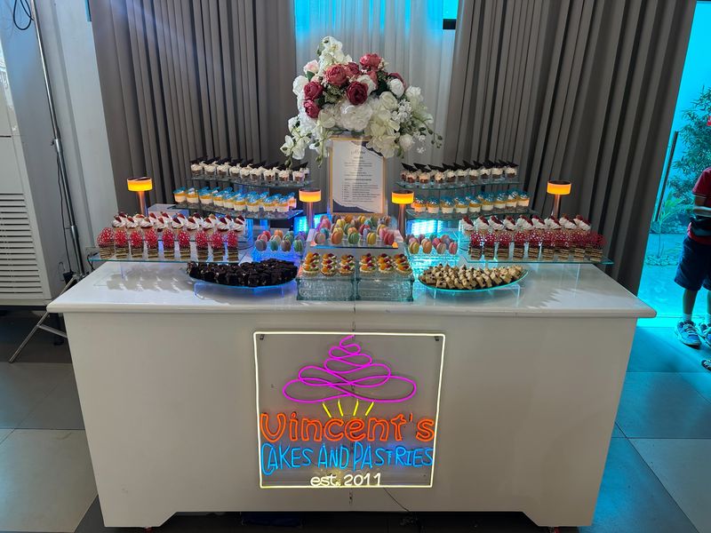 Dessert table with rows of mini cakes, dessert cups and pastries, a white floral centerpiece and a lit Vincent's Cakes and Pastries neon sign on the front