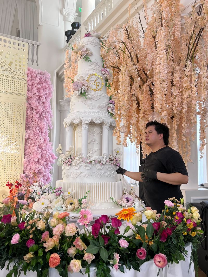 A cake artist in gloves adjusting decorations on a very tall white multi-tier wedding cake surrounded by fresh flowers at an event venue