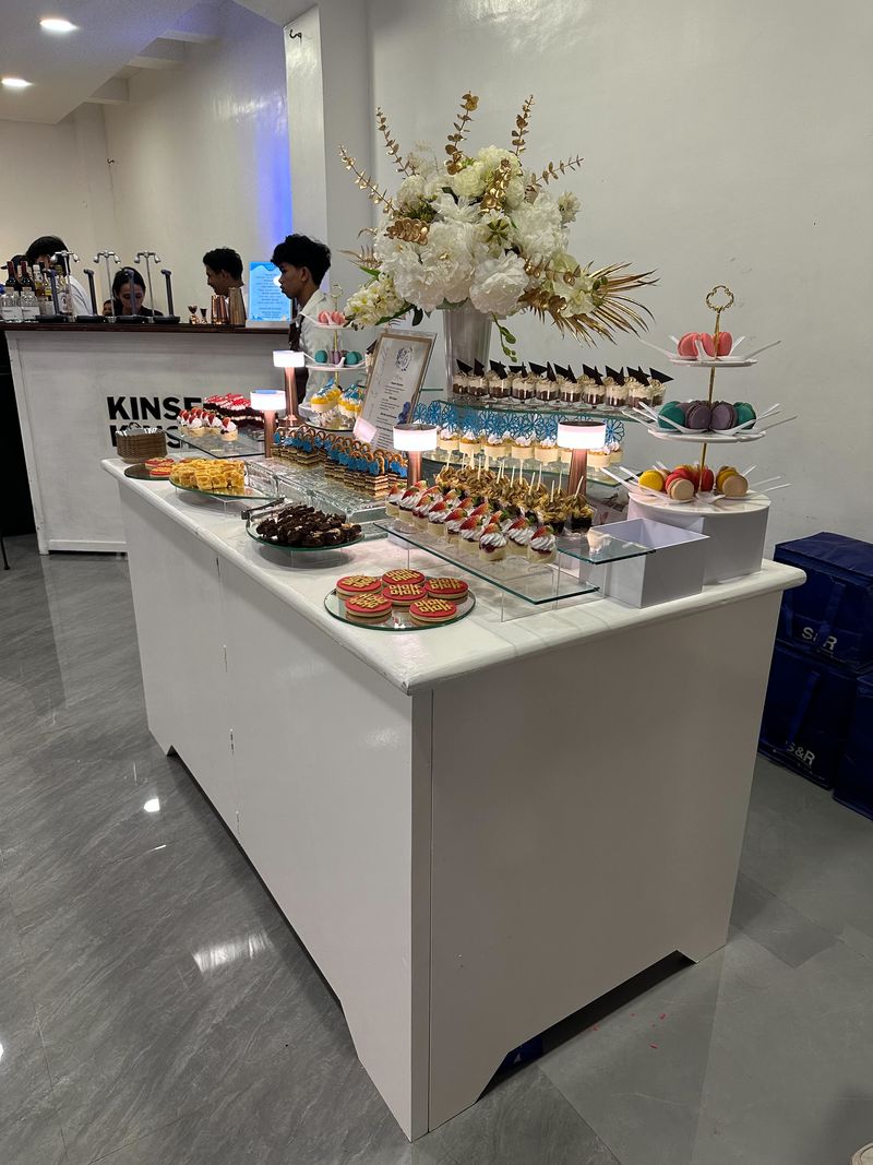 Full dessert buffet table with tiered stands of macarons, dessert cups, mini cakes and cookies under a large white and gold floral arrangement