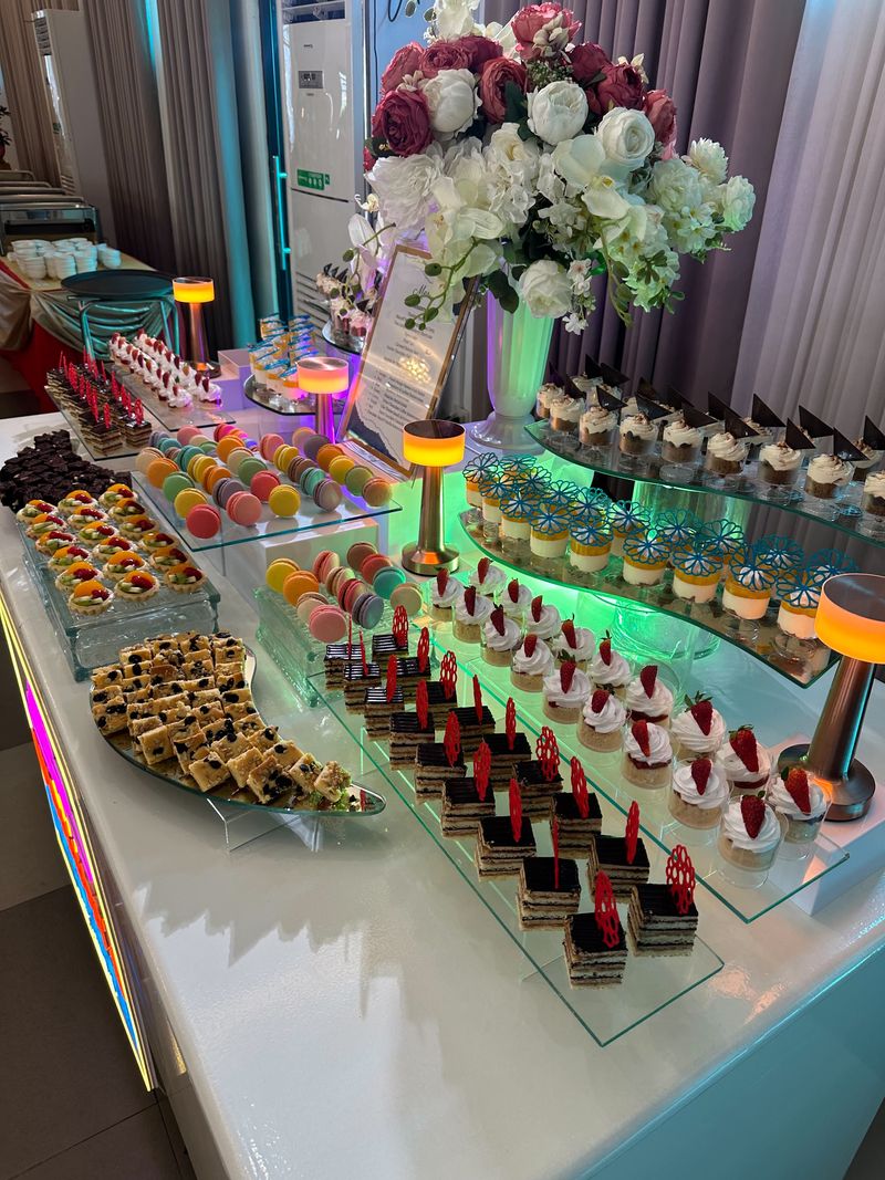 Dessert buffet on curved glass tiers with macarons, fruit tarts, layered mini cakes, strawberry-topped cups and a white and burgundy floral centerpiece
