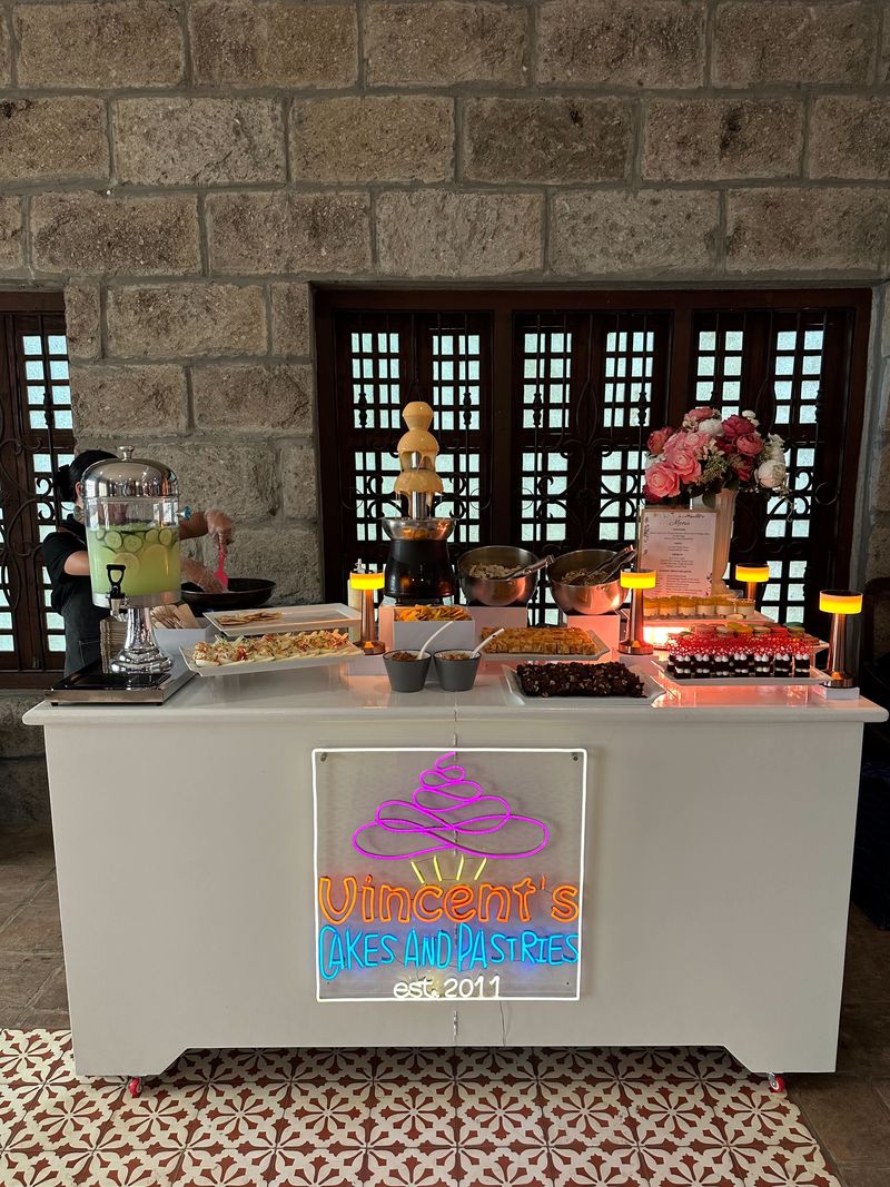Dessert station with a chocolate fountain, a drink dispenser, trays of treats and a Vincent's Cakes and Pastries neon sign against a stone wall