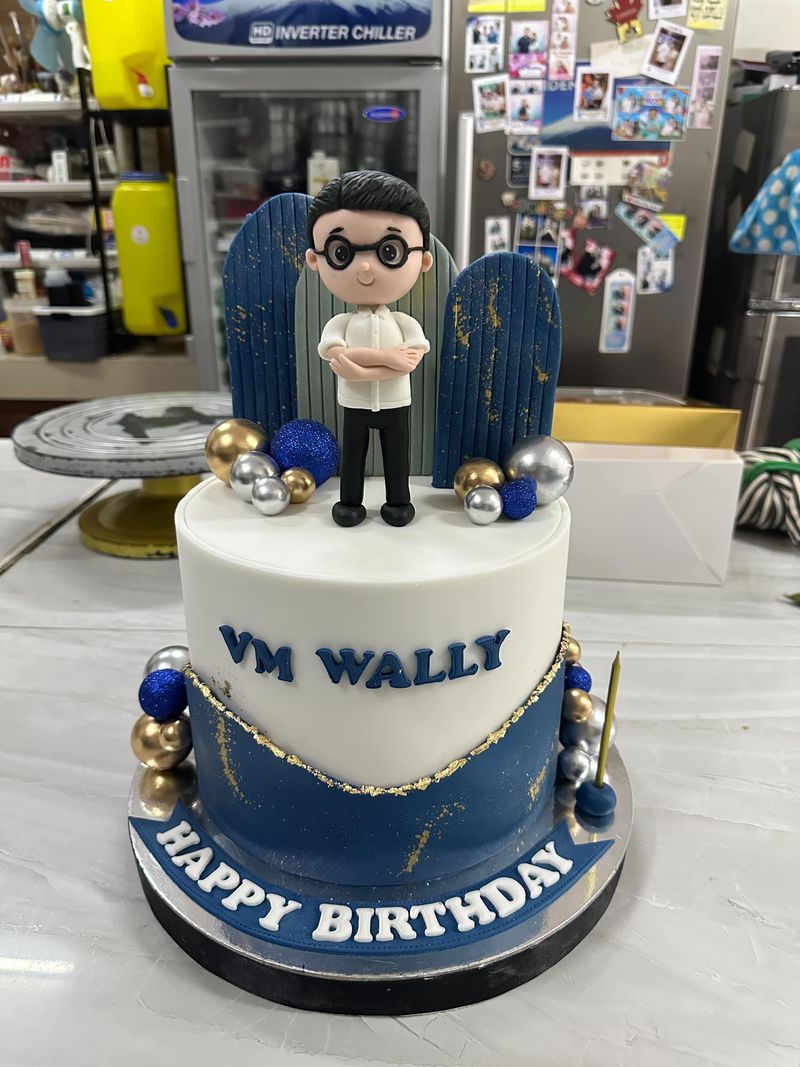 Two-tier white and navy birthday cake with a sculpted figurine topper, gold and blue spheres and a Happy Birthday banner