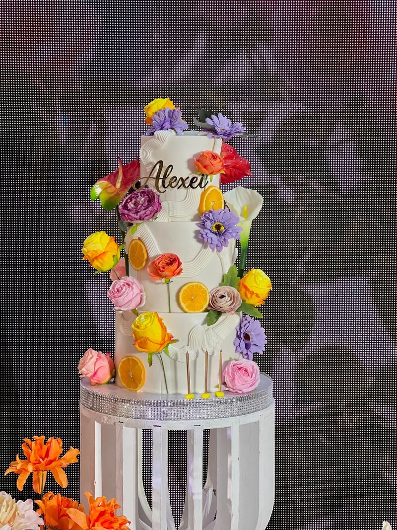 Three-tier white cake with a gold name topper, bright roses, gerberas, calla lilies and orange slices on a white stand