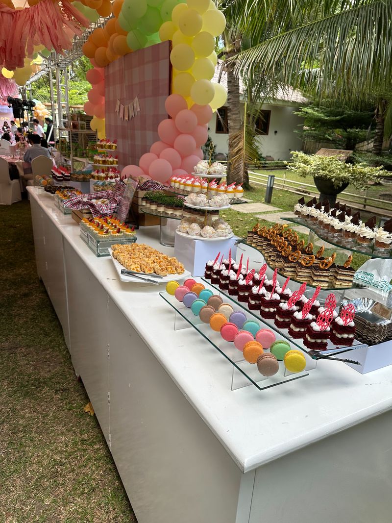 Long outdoor dessert table at a garden party with macarons, red mini cakes, cupcakes and bars in front of a pink gingham and balloon backdrop
