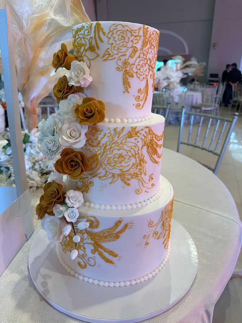 Three-tier white cake with gold lace-style detailing and a cascade of white and gold roses at an event venue