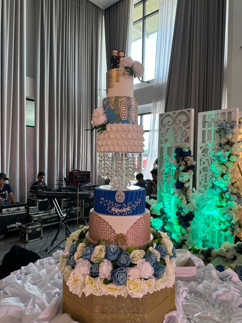 Towering multi-tier wedding cake in blue, gold, white and blush with a crystal-drop tier, a couple figurine topper and a base of blue and cream roses
