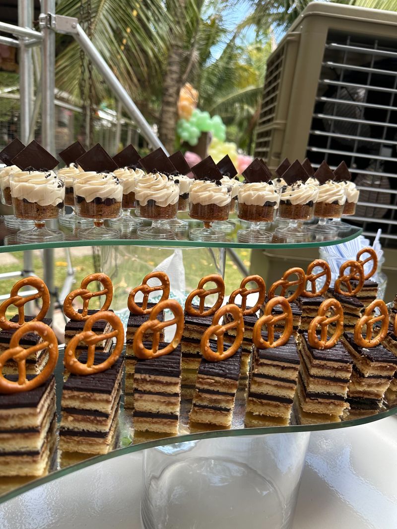 Layered chocolate-glazed mini cake slices topped with pretzels below a row of layered dessert cups with chocolate triangles