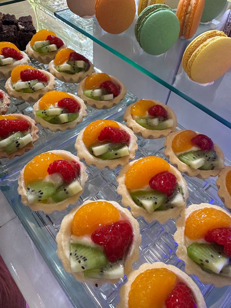 Mini fruit tarts topped with mandarin, strawberry and kiwi on a glass tray, with macarons on the tier above