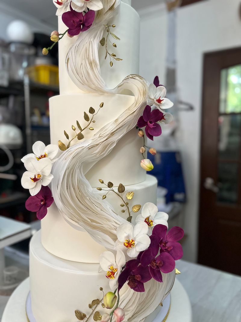 Close-up of a white tiered cake with a sculpted ivory ribbon, purple and white orchid decorations and gold leaf sprigs