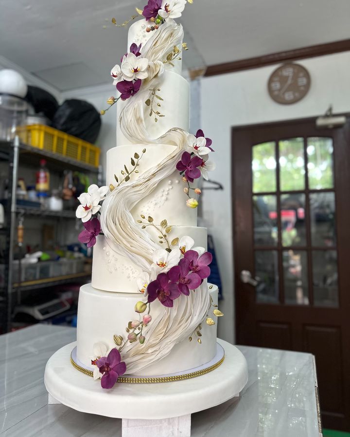 Tall multi-tier white cake with a flowing ivory ribbon sculpture, purple and white orchid decorations, gold leaf sprigs and pearl details