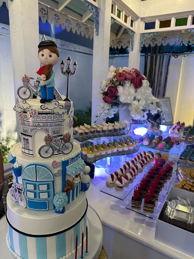 Three-tier Paris-themed cake with an Arc de Triomphe illustration, bicycles and a figurine topper, beside a dessert table of mini pastries