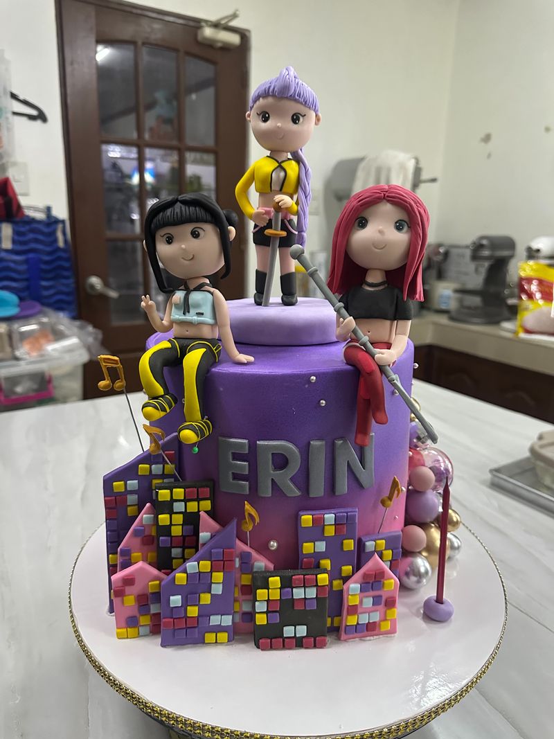 Purple birthday cake with three performer figurines, a microphone stand, music notes and a city-skyline base