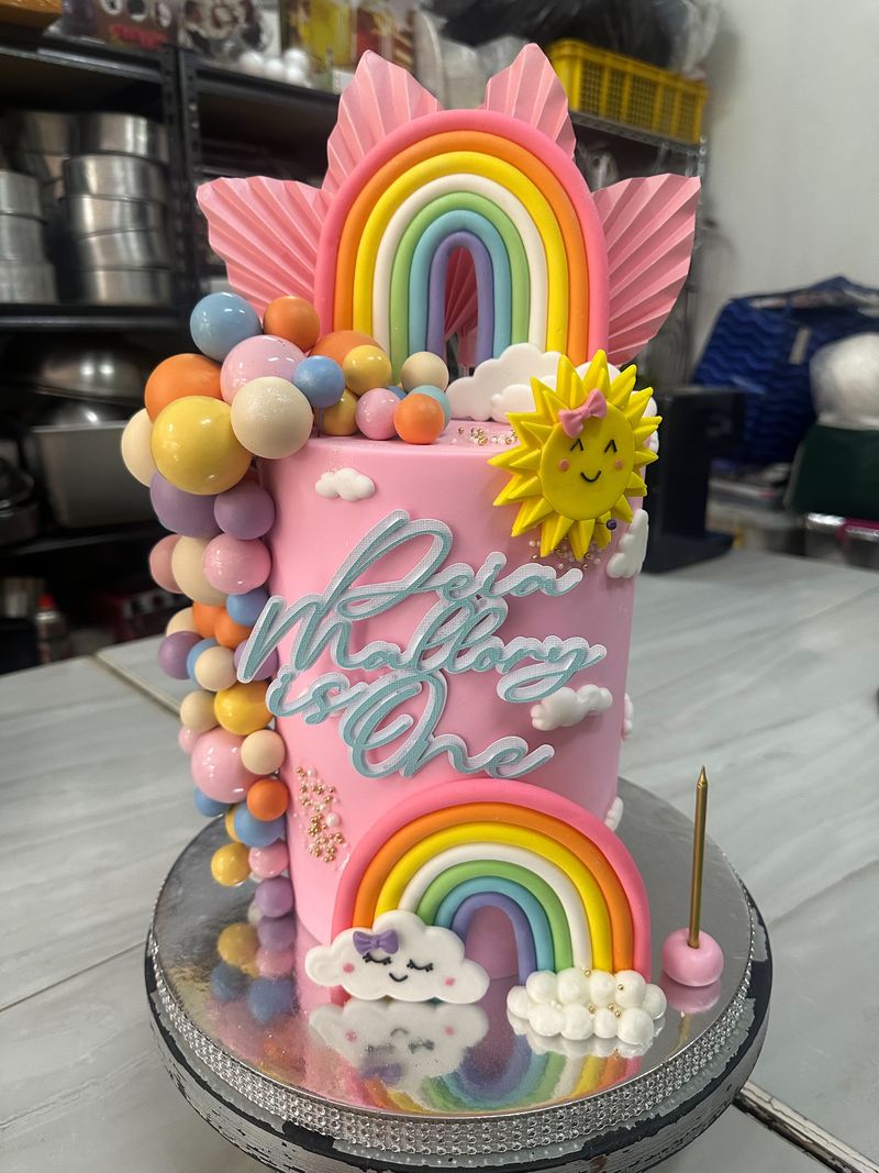 Tall pink first-birthday cake with rainbow, smiling sun and cloud decorations, pastel spheres and a script name topper