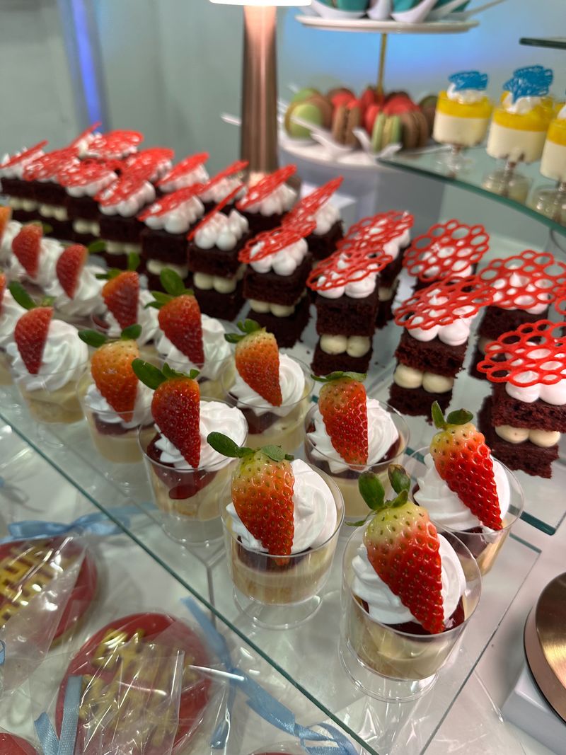 Dessert cups topped with whipped cream and fresh strawberries beside red layered mini cakes on glass tiers