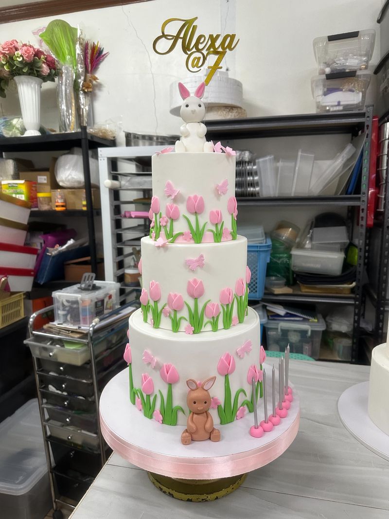 Three-tier white cake decorated with pink tulips, butterflies and bunny figurines, with a gold number-seven topper