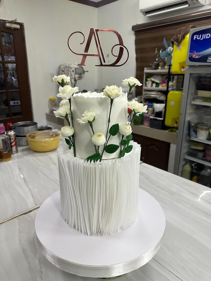 Two-tier white cake with a pleated bottom tier, white rose stems climbing the top tier and a rose-gold monogram topper